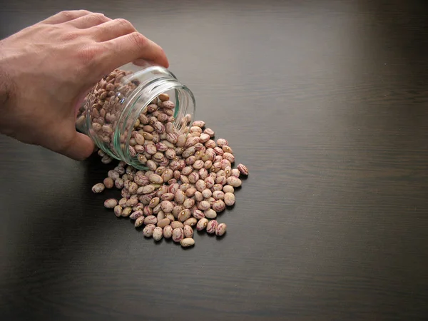 Pinto bean seeds hand dish.Pinto bean seeds are poured from a glass jar on brown wooden surface.Closeup of dried bean seeds in a glass jar on wooden table. In a glass dish. Hand on a glass jar. Background