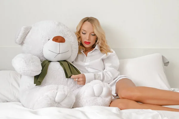 Ein junges Mädchen im weißen Hemd auf dem Bett spielt mit einem weißen großen Bärenspielzeug. — Stockfoto