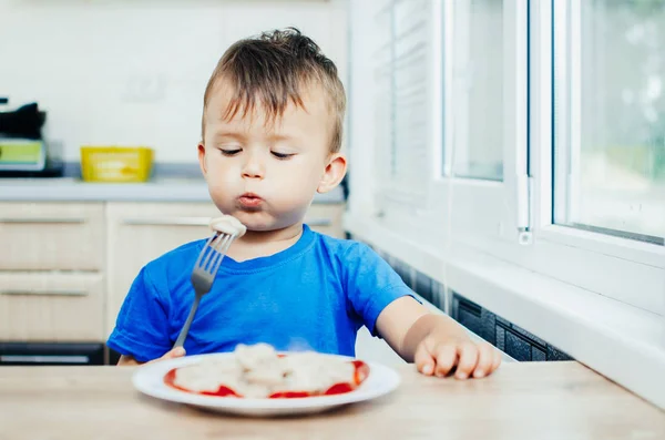 Hungry baby eating dumplings in the kitchen sitting on a chair in a blue t-shirt