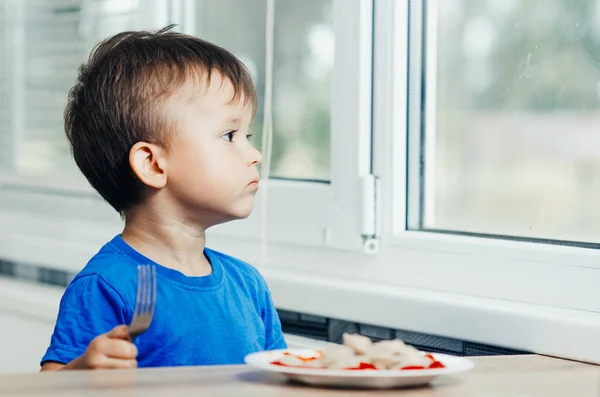 Hungry baby eating dumplings in the kitchen sitting on a chair in a blue t-shirt