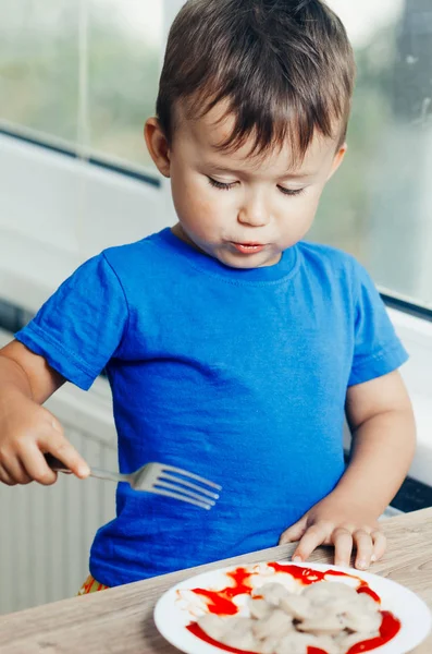 Hungry baby eating dumplings in the kitchen sitting on a chair in a blue t-shirt