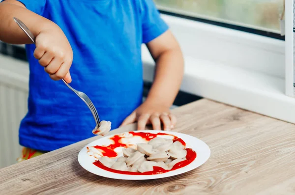 Hungry baby eating dumplings in the kitchen sitting on a chair in a blue t-shirt