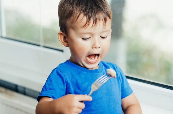 Hungry baby eating dumplings in the kitchen sitting on a chair in a blue t-shirt