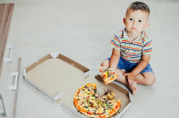 the child sits on the floor and eats pizza very appetizing and greedy, in shorts and a t-shirt