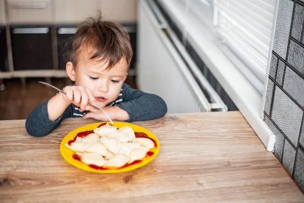 Hungry child eating dumplings in the kitchen, sitting at the table in a gray jacket