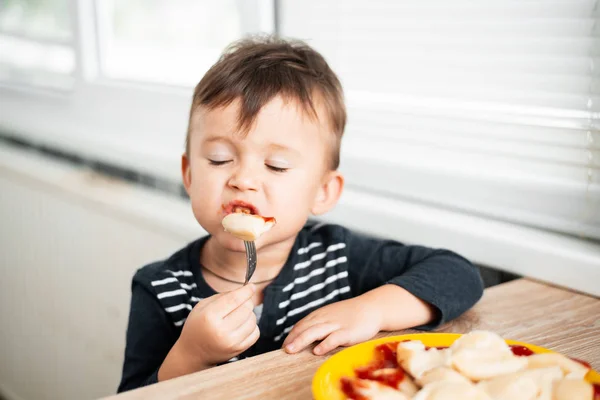 Hungry child eating dumplings in the kitchen, sitting at the table in a gray jacket