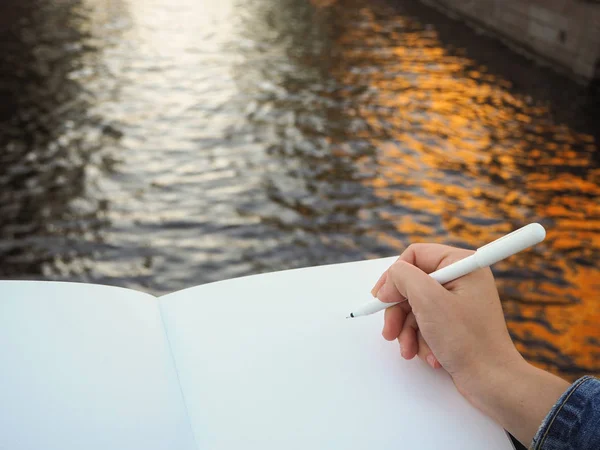 Kişinin elini boş beyaz defter tutan kendi fikirlerini yazmaya hazırlanıyor mockup. Bir kalem tutan kadın el yakın ve su arka plan üzerinde eskiz defteri sayfaları açtı. Düşüncelerinizi veya hedeflerinizi yazın.