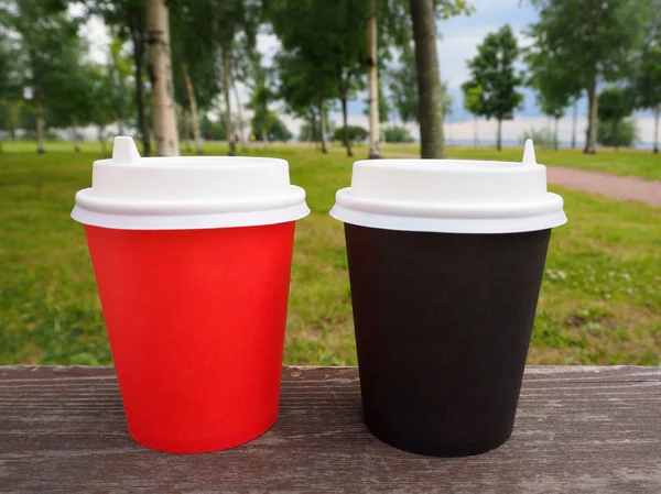 Mockup of red and brown paper takeaway coffee cups on wooden surface on natural summer park background, for product display montage. Close-up view of disposable paper cup with plastic lid. Polystyrene coffee drinking mug mock up front view. Clear pla