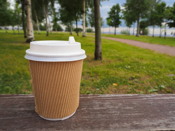 Mockup of brown paper takeaway coffee cup on wooden surface on summer park background, for product display montage. Close-up view of disposable paper cup with plastic lid. Clear plain tea take away package.