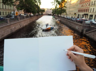 Kişinin elini boş beyaz defter tutan kendi fikirlerini yazmaya hazırlanıyor mockup. Bir kalem tutan kadın el yakın ve şehir kanalında yüzen teknenin arka planda eskiz defteri sayfaları açıldı. 