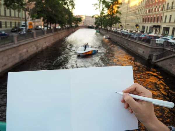 Kişinin elini boş beyaz defter tutan kendi fikirlerini yazmaya hazırlanıyor mockup. Bir kalem tutan kadın el yakın ve şehir kanalında yüzen teknenin arka planda eskiz defteri sayfaları açıldı. 