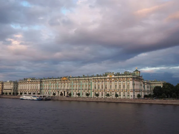 The State Hermitage Museum in St. Petersburg, with a touristic boat in front of it, on the background of stormy different color sky and dark water of Neva river. Panoramic view of the Winter Palace building on the Palace Embankment.