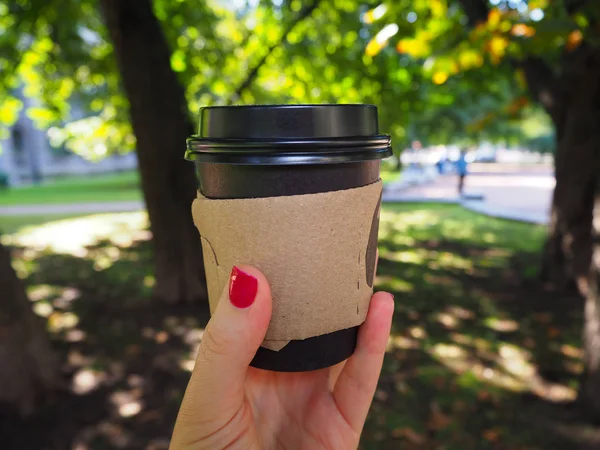 Disposable black paper coffee cup in woman\'s hand with red manicure on the park background. Eco friendly lifestyle. Coffeine addiction problem. Go Green, Zero waste, Save the planet, Earth day, Plastic Free, Recycle concept.