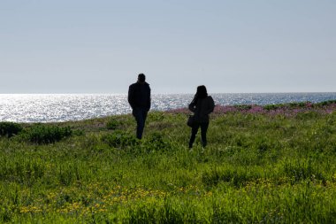 Denizin arka planında arka ışıkta birkaç kişi çimenlerin üzerinde yürüyor.