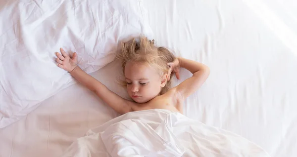Χαριτωμένο μικρό ξανθό παιδί ξυπνά μετά τον ύπνο σε λευκά σεντόνια. Εικόνα Αρχείου