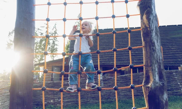Симпатична блондинка біла дитина піднімається вгору по мережі на дитячому майданчику зовні — стокове фото
