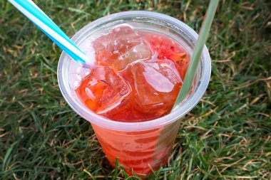 Kırmızı sangria, plastik bir bardakta buz ve çimenlerin üzerinde saman. Buzlu limonata, çimenlikte bir bardak serinletici bir içecek bir üst görünümü.