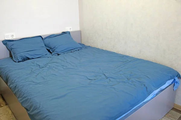 A large gray double bed is in the corner of a light gray room. The bed is covered with monochromatic blue linen, two pillows on the bed and two sockets on the wall. Crumpled empty bed in the morning.