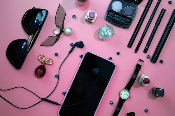 Women\'s cosmetics and accessories top view. Composition of a phone, watch, headphones and cosmetics on a pink background. What is in a woman\'s purse.