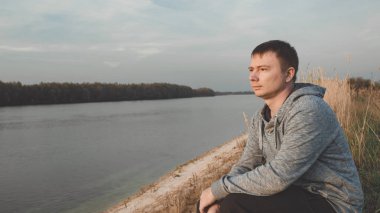 Adam nehir kıyısında oturuyor ve düşünceli bir şekilde batan güneşe bakıyor. Akşamüstü nehir kenarında.