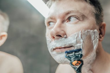 Adam banyo aynasının yanında tıraş oluyor. Elinde bir jiletle