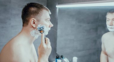 Adam banyo aynasının yanında tıraş oluyor. Elinde bir jiletle