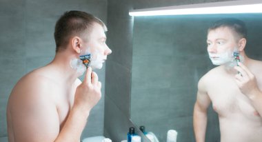 Adam banyo aynasının yanında tıraş oluyor. Elinde bir jiletle