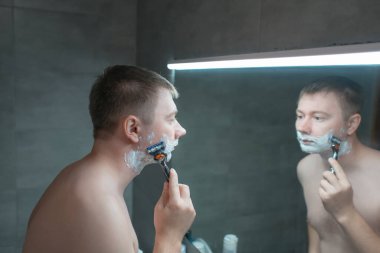 Adam banyo aynasının yanında tıraş oluyor. Elinde bir jiletle