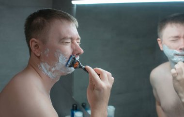 Adam banyo aynasının yanında tıraş oluyor. Elinde bir jiletle