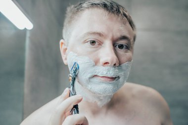 Adam banyo aynasının yanında tıraş oluyor. Elinde bir jiletle