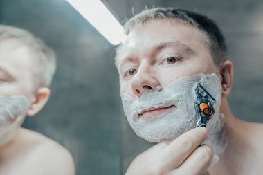 Adam banyo aynasının yanında tıraş oluyor. Elinde bir jiletle