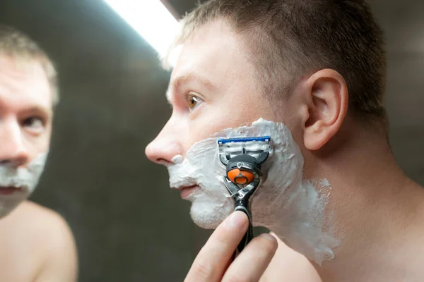 那家伙在浴室镜子旁边刮胡子 手里拿着剃须刀 — 图库照片