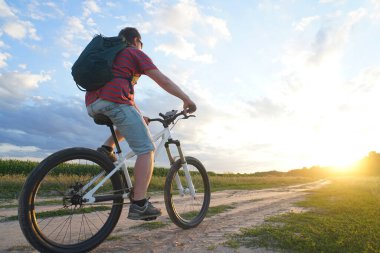 Dağ bisikletli bir adam gün batımında toprak bir yola çıkıyor.