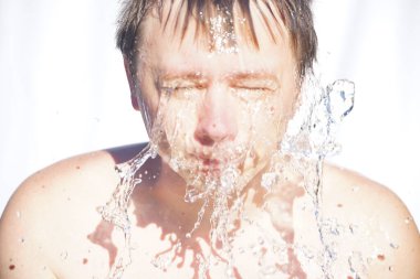 Adam yüzünü suyla yıkıyor, damlalar farklı yönlere uçuyor, beyaz bir arka planda.