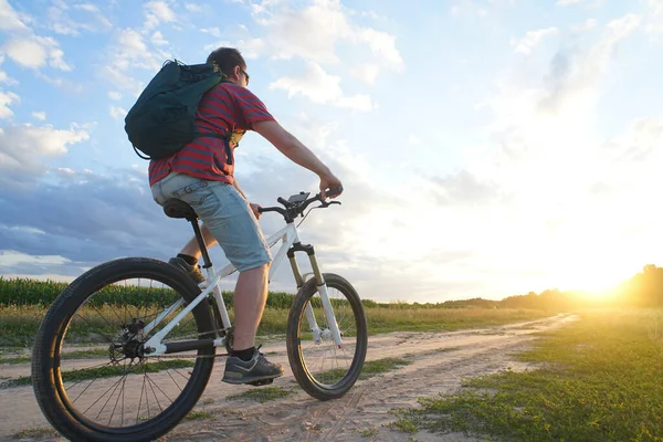 Dağ bisikletli bir adam gün batımında toprak bir yola çıkıyor.