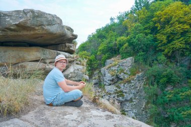 Bir adam dağlarda bir kayanın üzerinde oturuyor, bir taşın üzerinde, tişört ve şort giymiş.