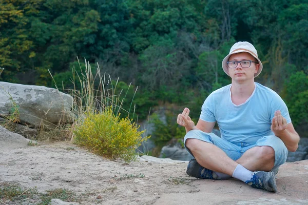 Bir adam dağlarda bir kayanın üzerinde oturuyor, bir taşın üzerinde, tişört ve şort giymiş.