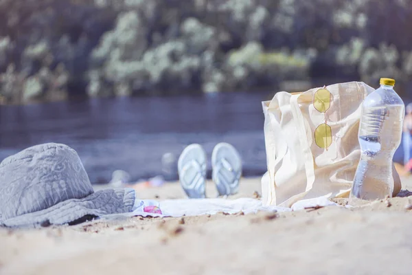 A light eco bag stands on a caved shore, at a sledge, near a towel and things. They weigh glasses on a bag and a bottle of water is standing nearby. Resting-place