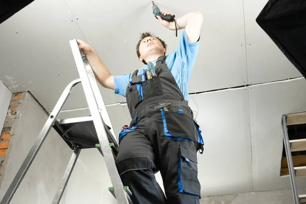 İş kıyafetleri içinde elinde elektrikli tornavidayla merdivenlerde duran bir adam. Evdeki yenileme çalışmalarına karşı.