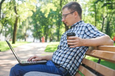 Dizüstü bilgisayarı olan bir adam parktaki bankta oturuyor. Elinde bir kupa kahve tutuyordu.