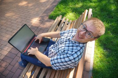 Adam parkta bir bankta oturuyor ve dizüstü bilgisayarda çalışıyor. Gülen surat.