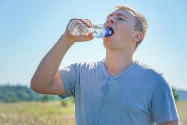 Gözlüklü ve tişörtlü bir adam elinde plastik bir su şişesi tutuyor.
