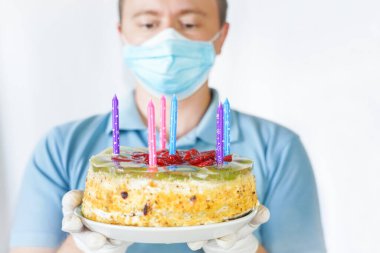 Maskeli ve eldivenli bir adam elinde pasta tutuyor. Covid-19 karantinası sırasında doğum günü kutlaması.