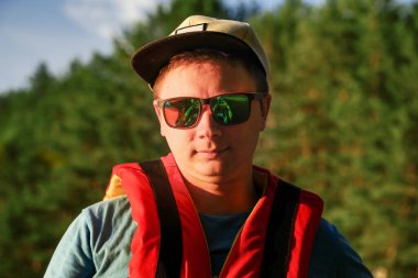 Can yeleği giymiş, gözlüklü ve arka planda şapka takmış bir adamın portresi..