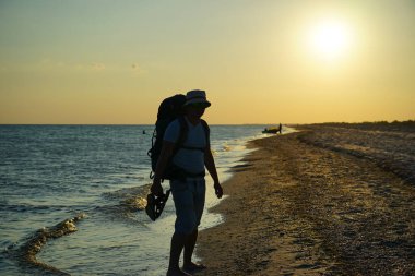 Sırt çantalı bir adamın siluetleri kumsalda yalınayak yürüyen, elinde sandalet tutan bir adamın.