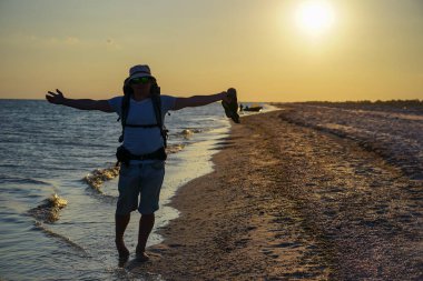 Şort, Panama tişörtü ve gözlüklü, büyük sırt çantalı bir turist gün batımında deniz kıyısında ıslak kum boyunca yürür..