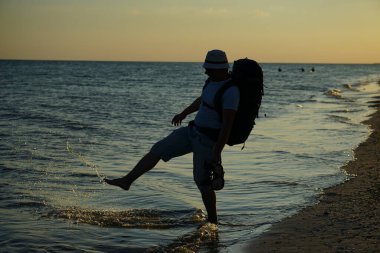 Şort, Panama tişörtü ve gözlüklü, büyük sırt çantalı bir turist gün batımında deniz kıyısında ıslak kum boyunca yürür..