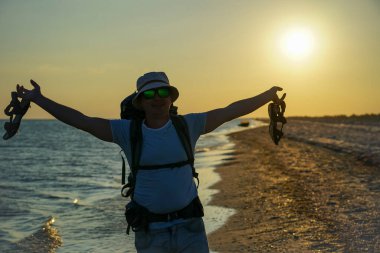 Adam akşamları deniz kıyısında ayakta duruyor ve elinde sandaletlerle geziyor. Büyük bir sırt çantasının arkasında, panama şapkası ve güneş gözlüğü takıyor.