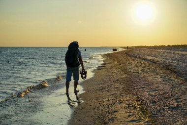 Sırt çantalı bir adam kumsalda yalınayak yürüyor, elinde sandaletleri var.
