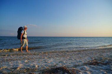 Sırt çantalı bir adam kumsalda yalınayak yürüyor, elinde sandaletleri var.